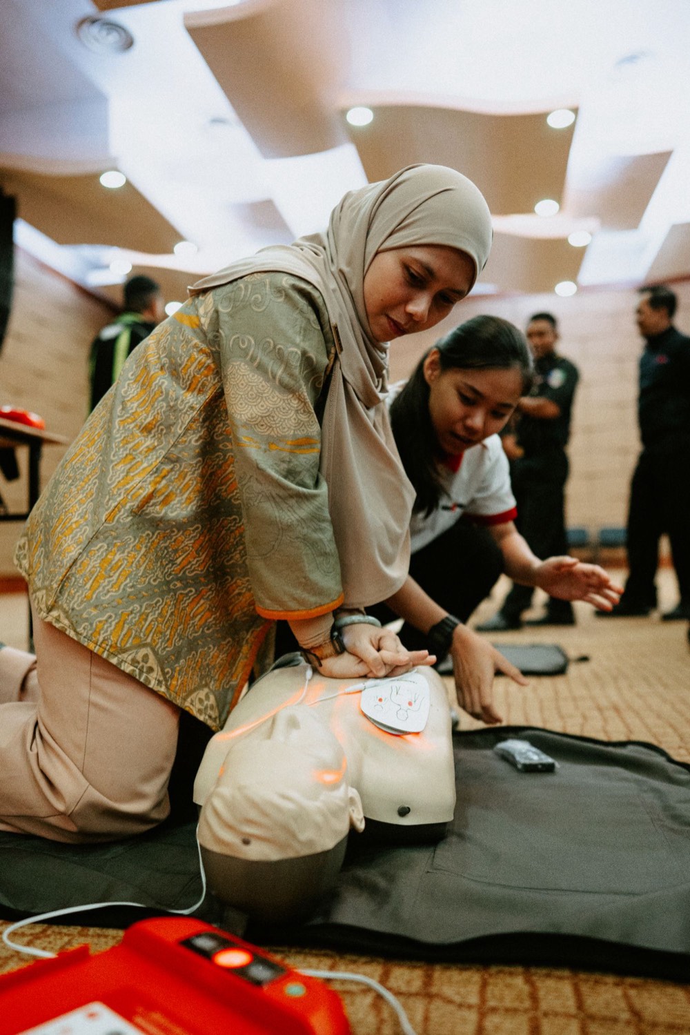 A MediShield trainer guiding a participant through chest compressions beside an AED trainer unit